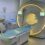 MRI scanner in a hospital room, showing a patient table in front of the machine, two monitors, and nearby medical equipment.