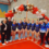 A group of gymnasts and coaches pose beneath a balloon arch in a gymnasium, all smiling at the camera.