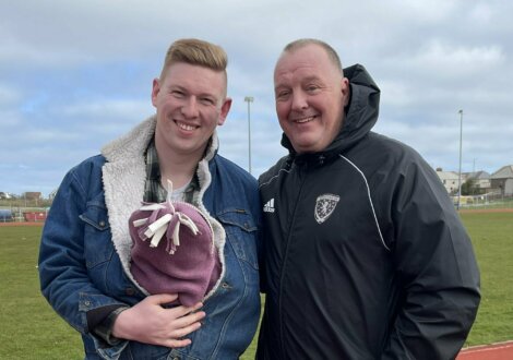 Two men stand on a running track, one holding a small child in a baby carrier. Both are smiling, and there is grass and houses in the background.