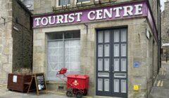 A stone building with a sign reading "TOURIST CENTRE," a grey door, a red cart, a wooden planter, and a blackboard sign on a paved street corner.