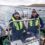 Two people wearing life jackets and knit hats operate a small motorboat on a body of water, with fish farm nets visible in the background.