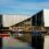 A modern angular building with large glass windows sits by a harbor, with several colorful small boats floating on the calm water in front.