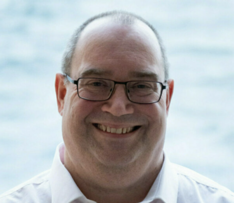 A middle-aged man with glasses, short hair, and a white collared shirt smiles at the camera with a blurred light blue background.