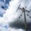 A helicopter carries a long pole suspended below it near a tall utility pole with power lines under a cloudy sky.