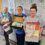 Three women stand in a hallway holding boxes filled with handmade baby clothes. Two are in scrubs, one in a coat. Behind them is a whiteboard with baby-themed drawings and notes.