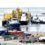 Several tugboats guide a large industrial vessel with yellow machinery at a busy dock. Warehouses, containers, and trucks are visible in the foreground.
