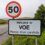 A speed limit sign of 50 stands above a roadside sign reading "Welcome to VOE. Please drive carefully," with greenery and hills in the background.
