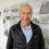 An older man in a black jacket stands smiling in front of a timeline display about the history and restoration of Swan LK243, with historical information and images visible on the wall.