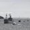 A convoy of ships, including a large gray vessel and smaller boats, sails near a lighthouse on a rocky shoreline under a cloudy sky.