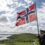 A uniformed man holds a large Norwegian flag on a hilltop with a rural landscape and cloudy sky in the background.