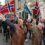 A group of people march down a street holding Norwegian flags and a blue banner, with spectators watching from the sidewalk.