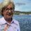 An older woman with short blonde hair and glasses stands by the waterfront, with boats and green hills visible across the water under a partly cloudy sky.