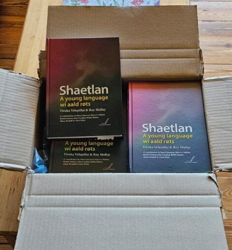 An open cardboard box containing multiple copies of the book "Shaetlan: A young language wi aald r&ograve;ts" by Viveka Velupillai and Roy Mullay.