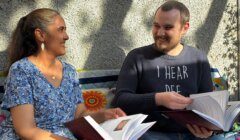 Two people sitting on a bench outdoors, smiling and holding open books, with sunlight casting shadows on the wall behind them.
