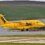 A yellow ADAC ambulance plane is landing on a runway near a body of water, with green hills in the background.