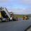 Construction workers operate heavy machinery to lay and smooth asphalt on a rural road under a cloudy sky, with vehicles nearby.
