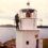 A person stands on the balcony of a white lighthouse overlooking a body of water with hills in the background, under an overcast sky.