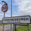 A road sign for Cunningsburgh with the message "Please Drive Carefully" stands beneath a circular 50 mph speed limit sign beside a rural road.