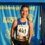 A young female runner wearing bib number 643 stands smiling in front of a Sri Chinmoy Races backdrop, with a medal around her neck.
