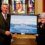 Two people stand indoors holding a framed painting of a ship at sea, with a stained glass window in the background.