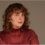 A person with curly brown hair wearing a textured maroon top looks at the camera against a neutral background.
