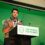 A person speaks at a podium with a sign reading "Scottish Greens. For People. For Planet." in front of a green backdrop.