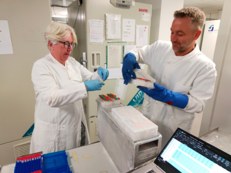 Two scientists in lab coats and blue gloves handle sample trays at a laboratory workstation, with a laptop displaying a spreadsheet nearby.