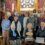 A group of people pose inside a church, some holding a wooden plaque and a certificate, with a priest standing at the front left.