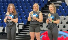 Three women stand on podium boxes holding awards in a sports arena with blue and white seats in the background.