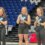 Three women stand on podium boxes holding awards in a sports arena with blue and white seats in the background.