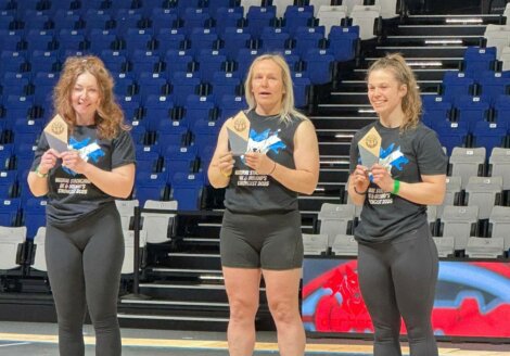 Three women stand on podium boxes holding awards in a sports arena with blue and white seats in the background.