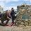 A man kneels to lay a wreath at a stone memorial surrounded by people during an outdoor remembrance ceremony.