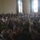 A large group of people sit closely together in rows inside a room with large windows, attentively facing in one direction.