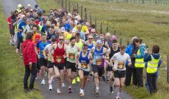 A group of runners at the starting line of a road race on a rural path, with marshals and spectators standing on the sidelines.