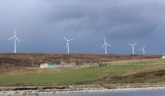 A row of wind turbines stands on a grassy hill under a cloudy sky, with a small building and stone ruins in the foreground near a body of water.