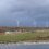 A row of wind turbines stands on a grassy hill under a cloudy sky, with a small building and stone ruins in the foreground near a body of water.