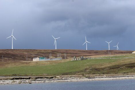 A row of wind turbines stands on a grassy hill under a cloudy sky, with a small building and stone ruins in the foreground near a body of water.