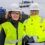 Two people wearing hard hats, safety glasses, and high-visibility jackets stand on a dock with a ship and water in the background.