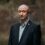 A middle-aged man with a shaved head in a dark suit jacket and light shirt stands outdoors against a blurred brown background.