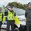Three police officers in uniform stand in front of a police vehicle parked outdoors on a cloudy day.