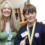 A woman and a boy stand side by side indoors. The boy wears a medal and a school sweatshirt, and both are smiling at the camera.