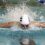 A swimmer wearing a swim cap and goggles performs the butterfly stroke in a pool, creating a splash of water behind them.