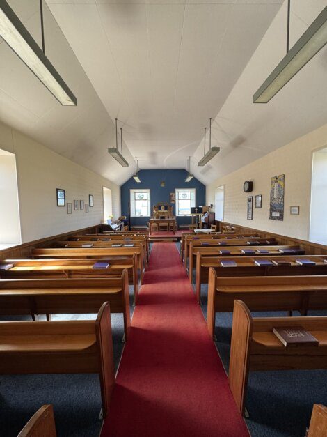Interior of a small church with wooden pews, red carpet aisle, and altar at the front, illuminated by natural light from side windows.