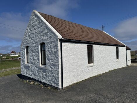 A small white stone building with a brown tiled roof, three windows, and an asphalt area, set in a rural area under a blue sky.