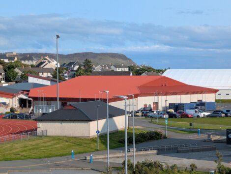 A sports complex with a red roof, parking lot, outdoor running track, and surrounding buildings, set against a hilly landscape.