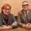 Two people with glasses, one wearing a scarf and green top, the other in a checked suit and tie, sit side by side at a wooden table against a beige wall.