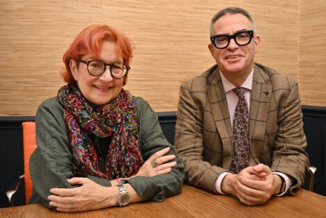 Two people with glasses, one wearing a scarf and green top, the other in a checked suit and tie, sit side by side at a wooden table against a beige wall.
