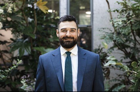 A bearded man in a blue suit and teal tie stands in front of leafy green plants, smiling at the camera.