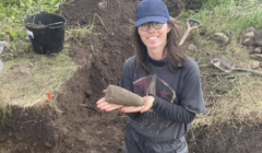 A person wearing glasses and a cap stands at an excavation site, holding a large, elongated stone artifact with both hands. Soil, tools, and a bucket are visible nearby.