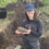 A person wearing glasses and a cap stands at an excavation site, holding a large, elongated stone artifact with both hands. Soil, tools, and a bucket are visible nearby.